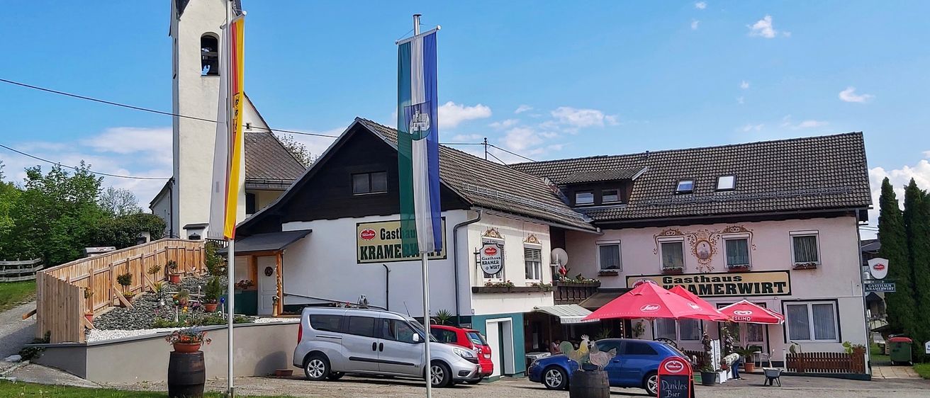 Ein deutsches Dorf mit einem Gasthaus Kramer vor einem Gebäude, hat geparkte Autos und Fahnen an der Seite.