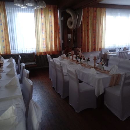 Ein Esszimmer mit weißen Stühlen und Tischen, die mit weißen Tüchern bedeckt sind. Es gibt Luftballons und Teddybären an der Wand und Blumen auf den Tischen.
