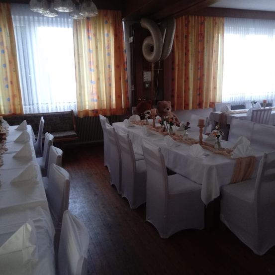 Ein Esszimmer mit weißen Stühlen und Tischen, die mit weißen Tüchern bedeckt sind. Es gibt Luftballons und Teddybären an der Wand und Blumen auf den Tischen.