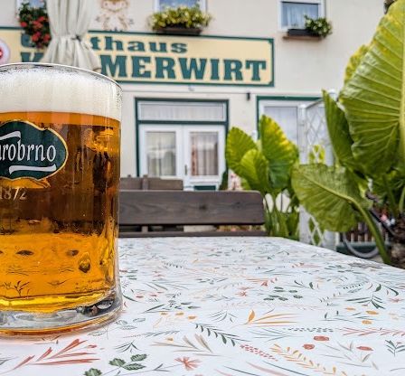 Ein Glas Bier mit einem Etikett, das 'StarobrnO 1872' liest, steht auf einem Tisch mit einer blumigen Tischdecke. Dahinter befindet sich ein Gebäude namens 'Haus MERWIR' mit Topfpflanzen an den Fenstern.