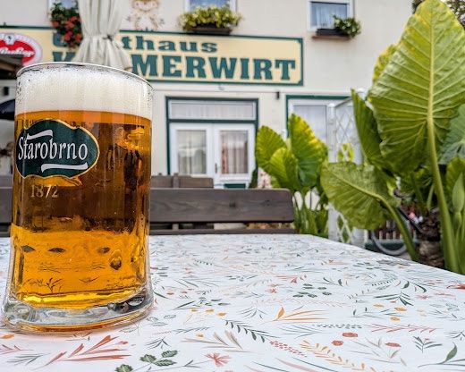 Ein Glas Bier mit einem Etikett, das 'StarobrnO 1872' liest, steht auf einem Tisch mit einer blumigen Tischdecke. Dahinter befindet sich ein Gebäude namens 'Haus MERWIR' mit Topfpflanzen an den Fenstern.
