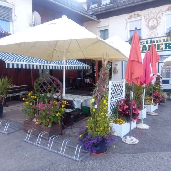 Eine Außensitzfläche mit Tischen und Stühlen unter Schirmen vor einem Gasthaus mit dem Schild Gasthaus Aver. Der Bereich ist mit Blumen in verschiedenen Farben und Topfpflanzen geschmückt.