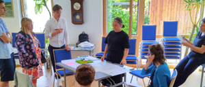 In einem Raum diskutiert eine Gruppe von Menschen, einige stehend, andere sitzend, wobei zwei Personen Papiere halten. Ein Tisch mit verschiedenen Gegenständen steht in der Mitte, und der Raum ist gut mit natürlichem Licht beleuchtet.