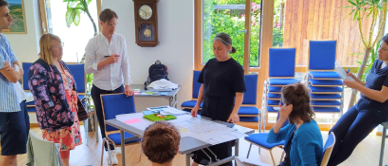 In einem Raum diskutiert eine Gruppe von Menschen, einige stehend, andere sitzend, wobei zwei Personen Papiere halten. Ein Tisch mit verschiedenen Gegenständen steht in der Mitte, und der Raum ist gut mit natürlichem Licht beleuchtet.