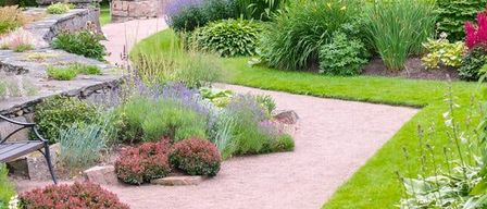 Ein Garten mit einem Kiesweg, Steinbänken und verschiedenen Pflanzen und Blumen. Bäume und Grün umgeben das Gebiet.