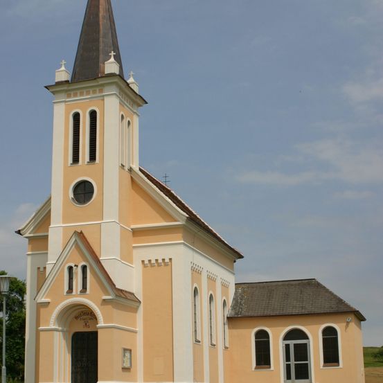 Eine beige Kirche mit einem spitzen Turm und einem braunen Dach. Die Fassade weist Bogenfenster, ein rundes Fenster und eine Holztür auf. Die Kirche hat einen kleinen Anbau mit einem Schieferdach.