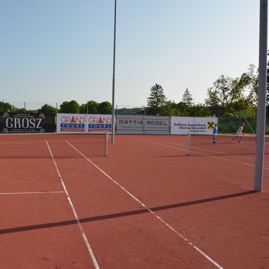 Ein Tennisplatz mit roter Oberfläche und weißen Markierungen. Es gibt Banner am Zaun. Zwei Personen spielen Tennis.