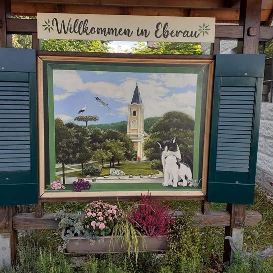 Ein gerahmtes Gemälde auf einem Holzständer zeigt eine Kirche, zwei Vögel und eine Katze. Das Gemälde trägt den Titel Willkommen in Eberau.