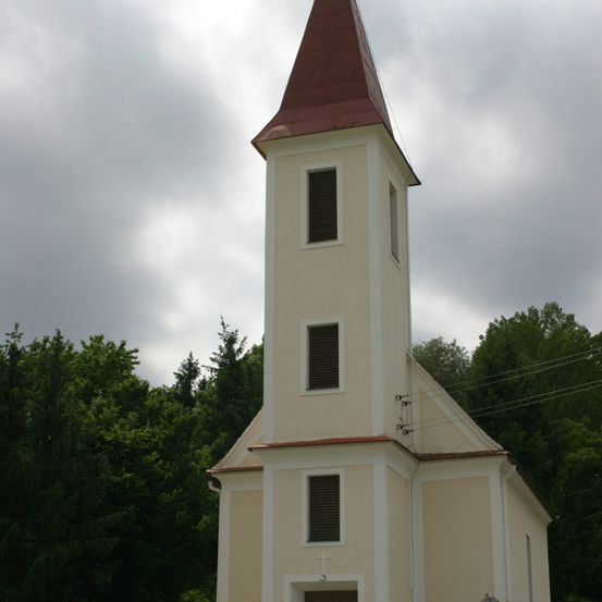 Eine weiße Kirche mit einem braunen Dach und Fenstern steht in einem ländlichen Gebiet bei bewölktem Himmel. Bäume umgeben die Kirche.