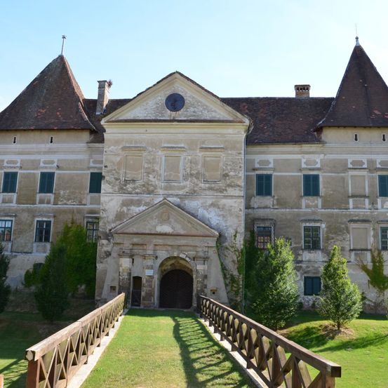 Ein altes Schloss mit braunen Dächern und grünen Fenstern, gelegen in einem Park. Es hat einen großen Eingang mit einem gewölbten Durchgang und einer Uhr darüber. Das Schloss ist von einem Rasen umgeben.