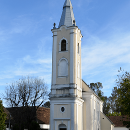 Eine Kirche mit einem hohen Turm steht gegen einen blauen Himmel. Der Turm hat gewölbte Fenster und ein spitzes Dach. Die Fassade der Kirche ist weiß mit einigen verwitterten Bereichen. In der Nähe befindet sich ein Baum mit spärlichen Blättern und ein braun gedecktes Gebäude.