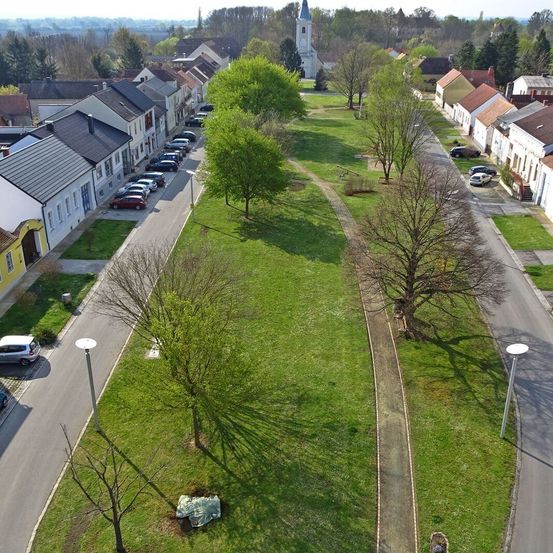 Luftaufnahme einer Straße mit geparkten Autos, einem Rasenfeld, Bäumen und einer Kirche in der Ferne.