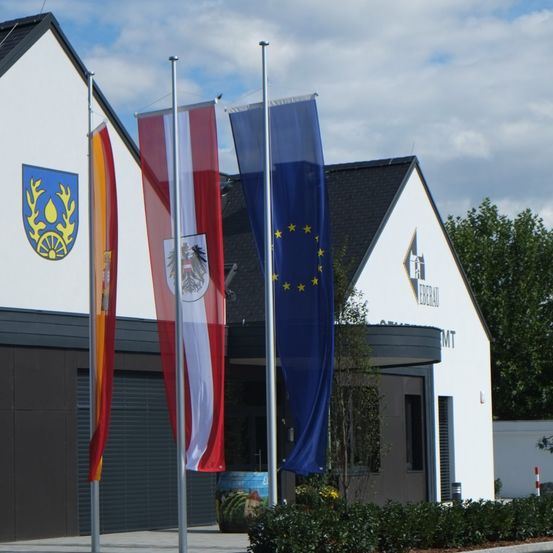 Ein Gebäude mit drei Fahnen davor: eine deutsche Flagge, eine Fahne mit einem Wappenschild und eine EU-Flagge. Das Gebäude hat ein weißes Äußeres mit einem schwarzen Dach.