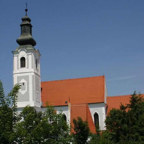 Ein hoher Kirchturm mit grünem Dach steht gegen einen blauen Himmel. Der Turm hat zwei Öffnungen auf beiden Seiten und ein kleines Fenster im oberen Teil. Die Kirche hat ein oranges Dach. Bäume umgeben das Gebäude.
