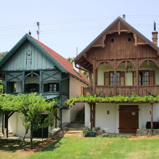 Zwei traditionelle Häuser nebeneinander, eines mit rotem Dach und grünen Akzenten, das andere mit brauner Holzfassade und grünen Reben auf dem Balkon. Beide haben hölzerne Balkone und Stufen, die zu den Eingängen führen.