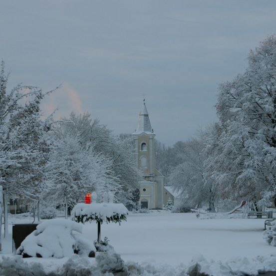 Eine schneebedeckte Landschaft zeigt eine Kirche mit einem hohen Turm, umgeben von schneebedeckten Bäumen. Schnee bedeckt den Boden und die nahen Pflanzen.