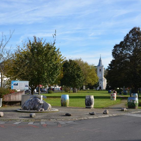 Ein großer Rasenplatz mit einem weißen Kirchturm im Hintergrund. Bunte Fässer und eine Steinskulptur stehen auf dem Bürgersteig. Bäume und Büsche umgeben das Gebiet.