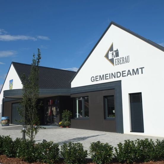 Die Vorderseite eines weißen Gebäudes mit schwarzem Dach, beschriftet mit 'Gemeindeamt Eberau'. Es hat Fenster, ein Logo und eine Pflanze davor.