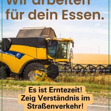 Ein gelber New Holland Mähdrescher erntet auf einem sonnigen Tag Getreide. Eine Person fährt den Mähdrescher. Der Bildtext lautet 'Wir arbeiten für dein Essen. Es ist Erntezeit! Zeige Verständnis im Straßenverkehr!'