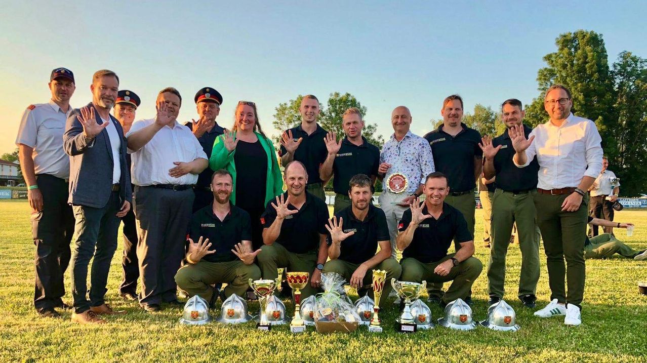 Eine Gruppe von Feuerwehrleuten in schwarzen Uniformen posiert für ein Foto, einige kniend und andere stehend, mit Trophäen und Geschenken vor ihnen an einem sonnigen Tag.