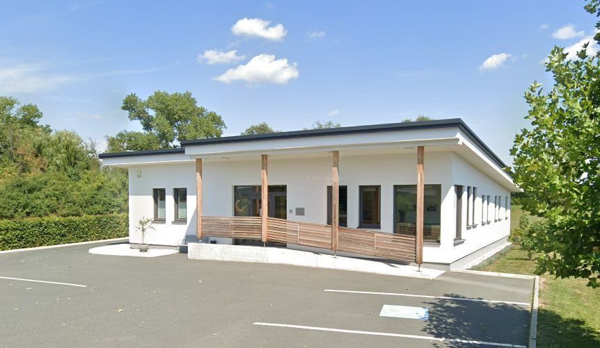 Ein modernes weißes Gebäude mit schwarzem Dach, hölzernem Vordach und mehreren Fenstern, in einer begrünten Umgebung mit blauem Himmel.