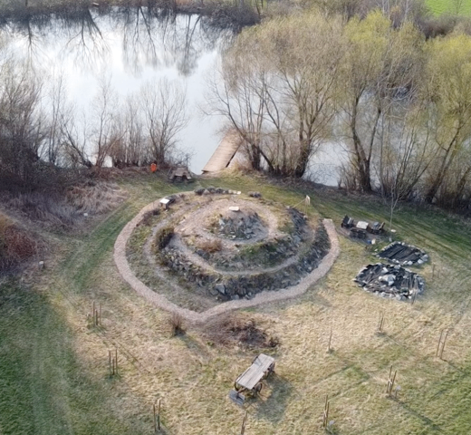 Aerial view of a grassy area with a spiral stone structure, near a water body, with a small wooden bridge and scattered trees.