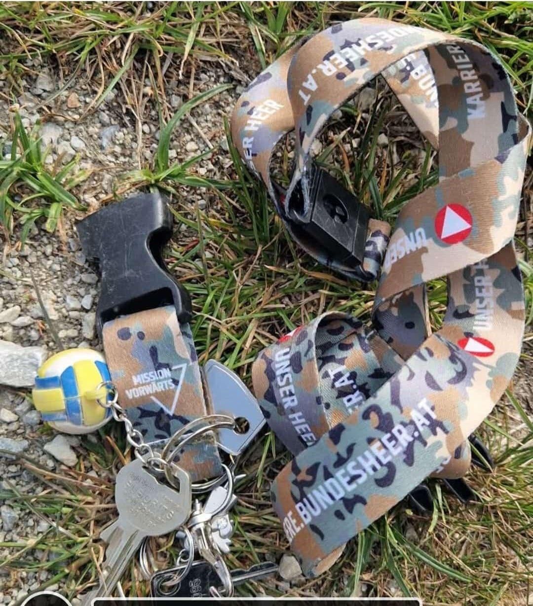 Camouflage lanyards, keys, and a ball are on grass. Lanyards say 'MISSION VORWAERTS' and 'WARRIOR'. The ball is yellow, blue, and white.