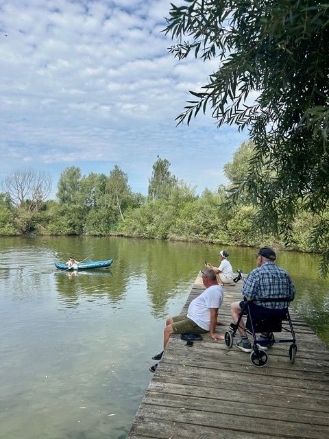 Drei Männer sitzen auf einem Holzsteg und beobachten eine Person in einem blauen Kajak auf einem Fluss. Der Fluss ist ruhig, und der Himmel ist blau mit Wolken.