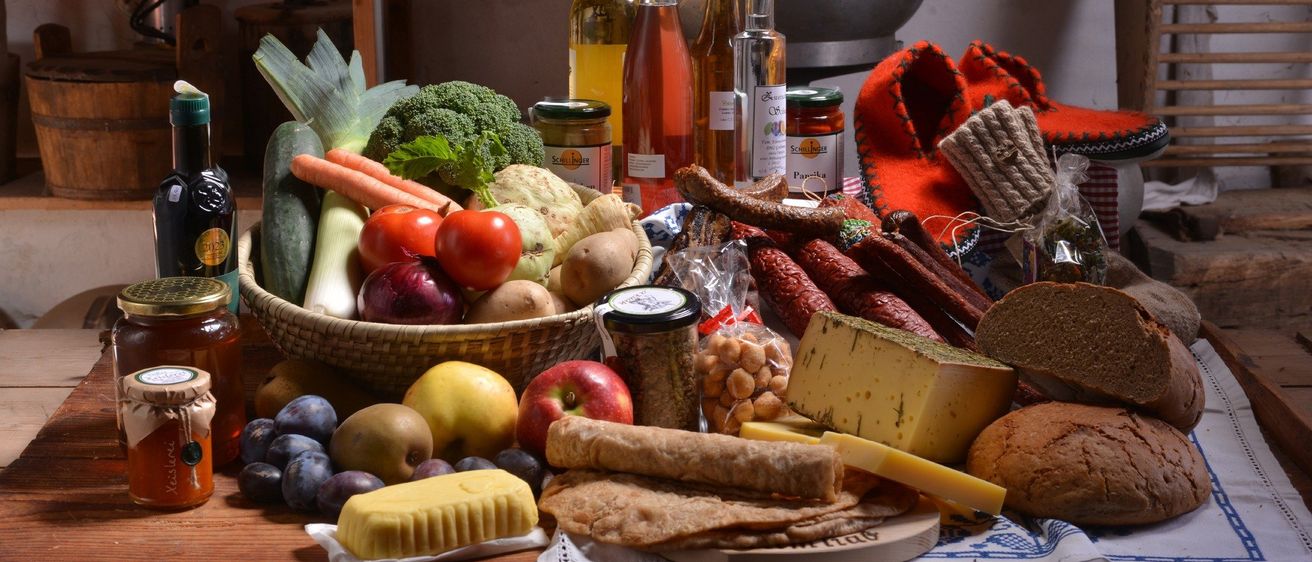 Ein Holztisch mit verschiedenen Lebensmitteln wie Gemüse, Obst, Fleisch, Käse und Brot. Flaschen mit Öl und Wein sind ebenfalls vorhanden.