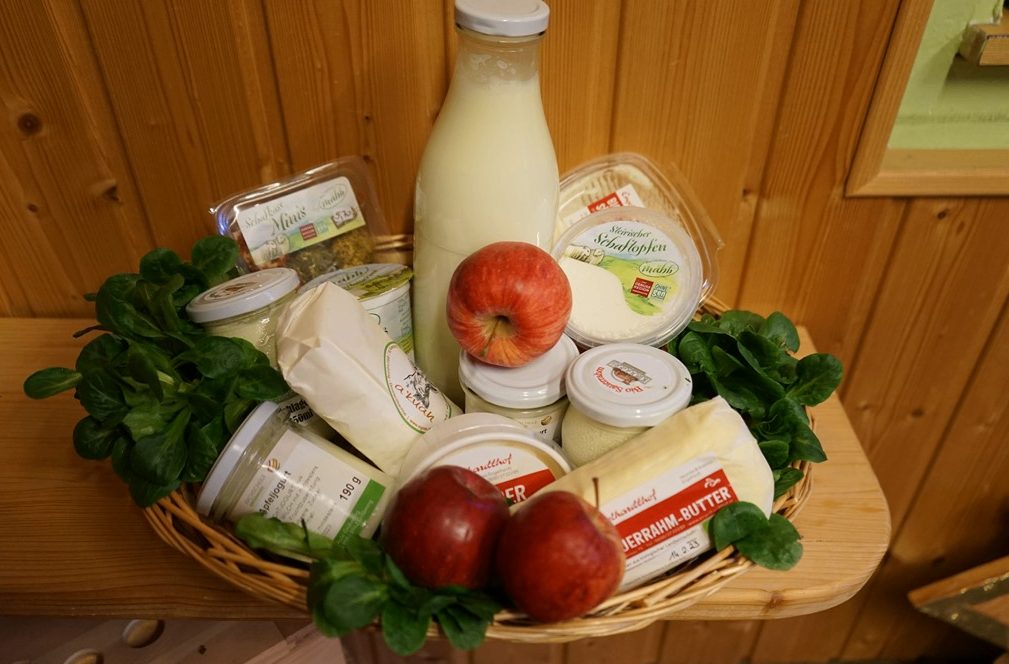 Ein Korb aus Weide gefüllt mit verschiedenen Milchprodukten, darunter eine Milchflasche, zwei Äpfel, Käse und Spinat. Die Artikel sind auf einem Holztisch angeordnet.