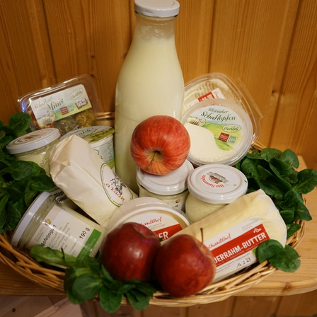 Ein Korb aus Weide gefüllt mit verschiedenen Milchprodukten, darunter eine Milchflasche, zwei Äpfel, Käse und Spinat. Die Artikel sind auf einem Holztisch angeordnet.