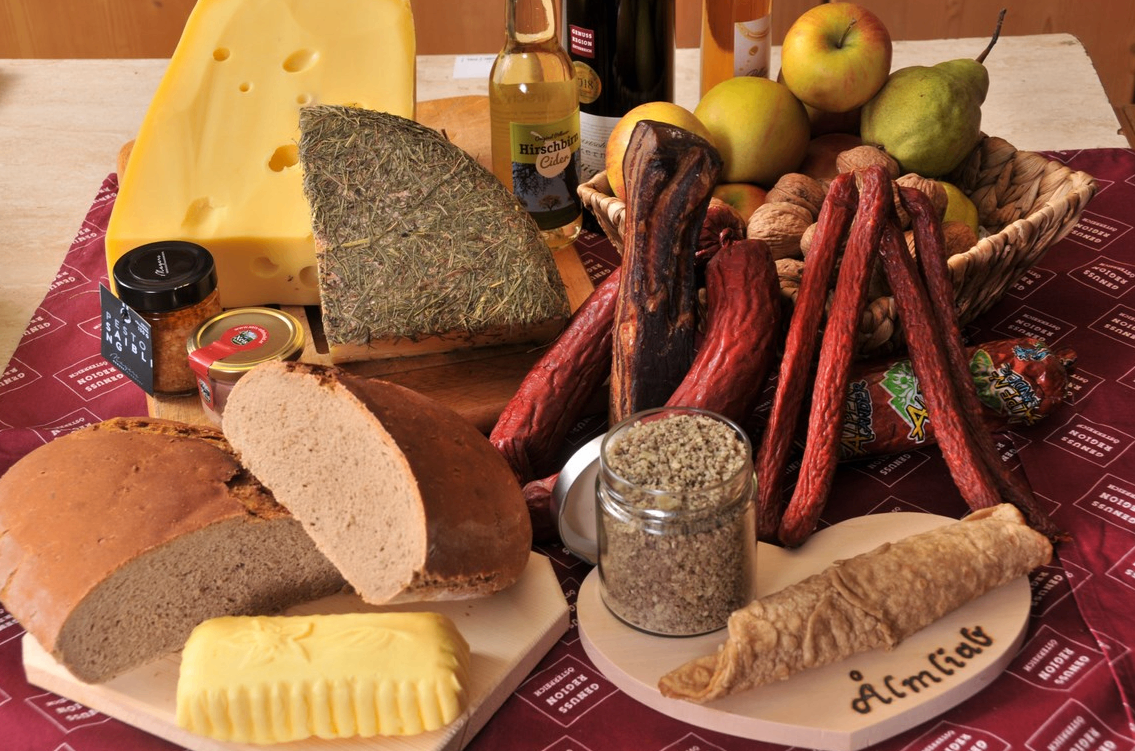 Eine Auswahl an Lebensmitteln ist ausgestellt, darunter Brot, Käse, Würste, Äpfel, Nüsse und ein Glas mit Gewürzen auf einem Tisch.