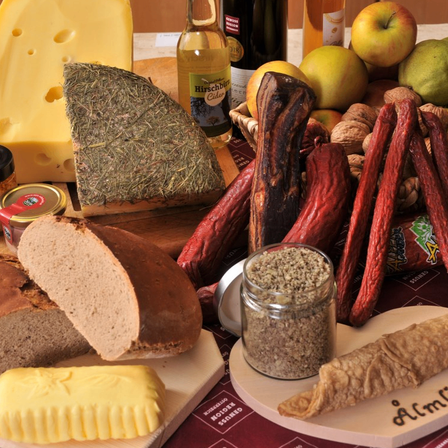 Eine Auswahl an Lebensmitteln ist ausgestellt, darunter Brot, Käse, Würste, Äpfel, Nüsse und ein Glas mit Gewürzen auf einem Tisch.
