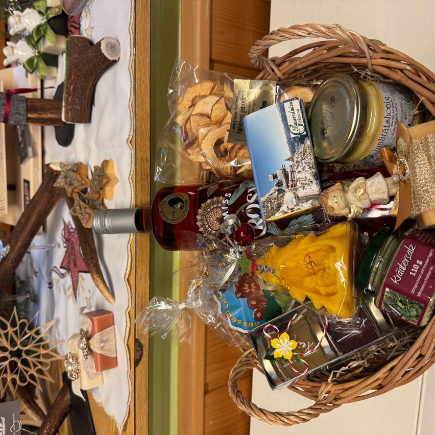 Ein Weidenkorb gefüllt mit verschiedenen Gegenständen, darunter eine Flasche Wein, verpackte Süßigkeiten und eine kleine Figur. Die Gegenstände sind ordentlich angeordnet, einige in Plastikverpackung. Der Korb steht auf einer Holzoberfläche.