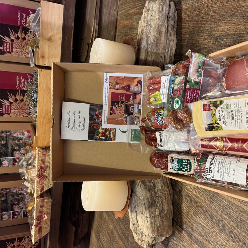 Eine braune Box ist geöffnet und zeigt eine Vielzahl verpackter Lebensmittel, einschließlich Würste, Käse und andere Artikel. Es gibt eine Karte mit Text. Hinter der Box befinden sich weitere Gegenstände, darunter ein Holzstamm, eine weiße Kerze und weitere Boxen.