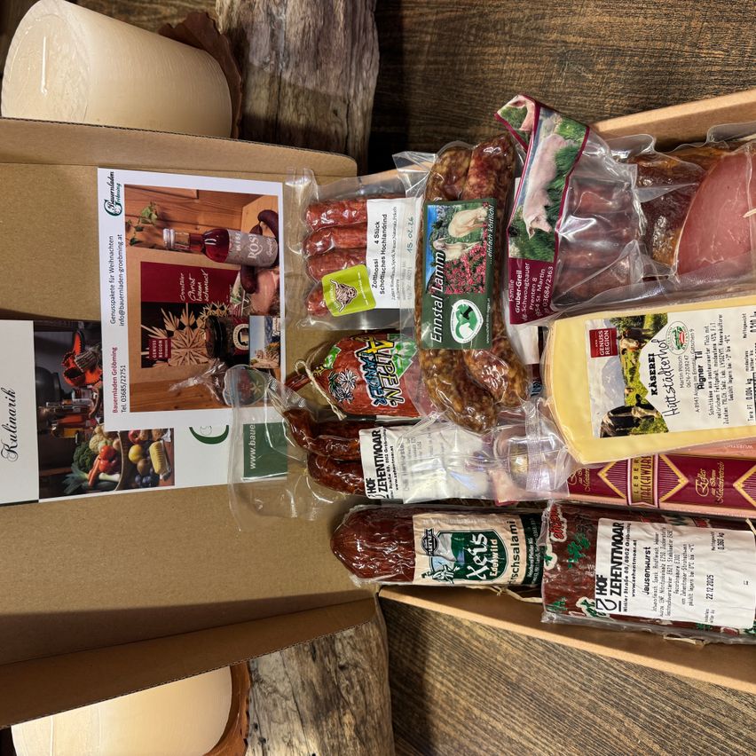 Ein brauner Karton mit einem Produktbild und verschiedenen vakuumverpackten Artikeln, darunter Würste, Salami und Käse, auf einer Holzoberfläche.