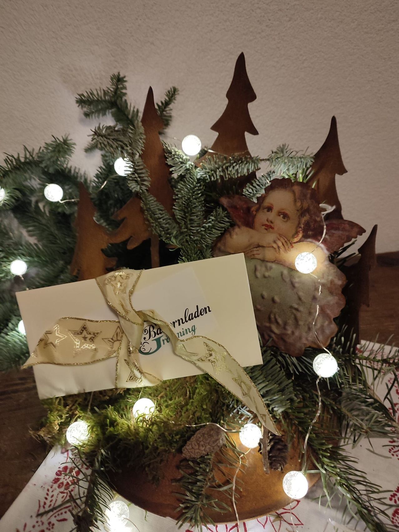Ein mit Lichtern geschmückter Weihnachtsbaum mit einer Engelsfigur, oben befindet sich eine Karte und ein Band mit der Aufschrift 'Barnladen'.
