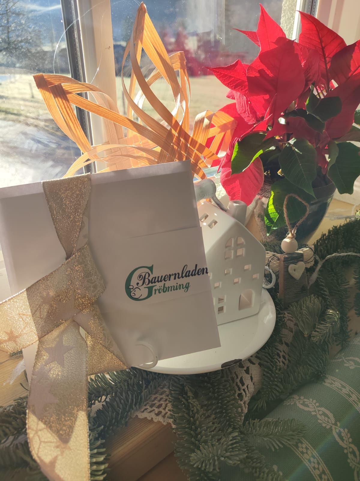 Eine Geschenkbox mit einer Schleife und dem Logo 'Bauernladen Grobming' ist in ein goldenes Band eingewickelt. Ein kleines Haus und eine Vase mit roten Weihnachtssternen stehen hinter dem Geschenk.