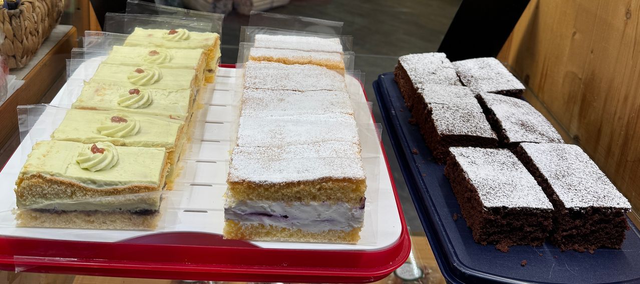 Eine Auswahl an Kuchen auf einem weißen und roten Tablett, mit einem Stück Kuchen mit Blaubeerfüllung und Puderzucker obenauf, neben einem blauen Tablett mit Brownies.