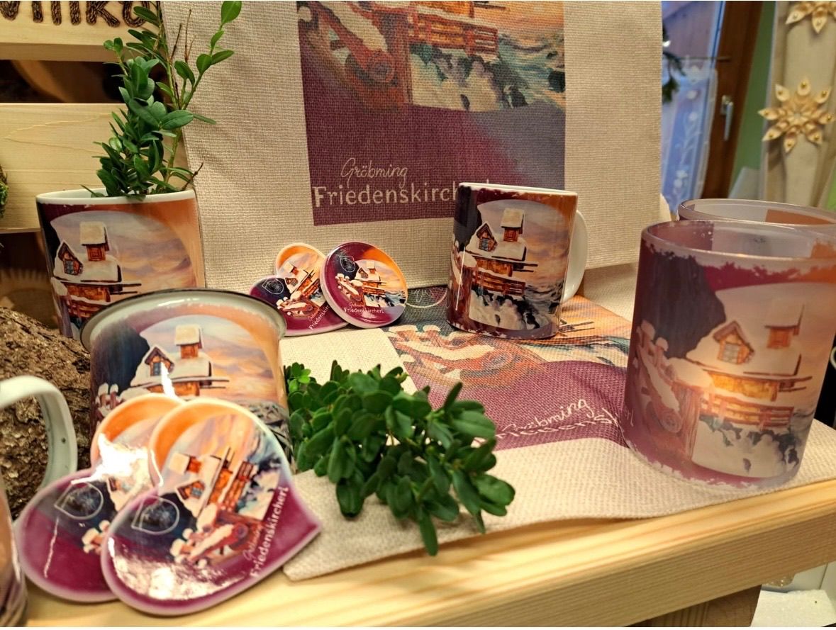 Ein Holztisch zeigt Tassen, Kerzen und herzförmige Kerzen mit Winterdorf-Bildern. Eine Pflanze und ein Schild mit 'Gröbming Friedenskirche' sind ebenfalls vorhanden.