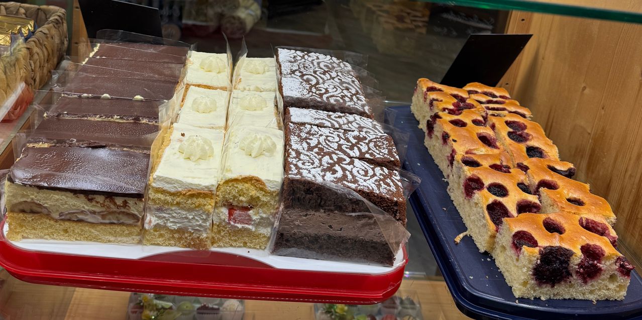 In einer Bäckerei sind mehrere Kuchen zur Schau gestellt. Die Kuchen umfassen Biskuitkuchen mit weißer Sahne, Schokoladenkuchen mit aufwendigen Mustern und eine Blaubeertarte. Sie stehen auf einem roten Tablett mit Plastikabdeckung.