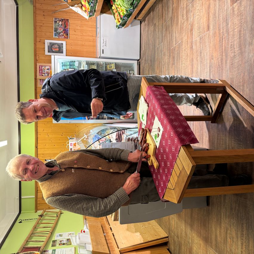 Zwei Männer sind drinnen, lächeln und schneiden Essen auf einem Tisch. Der Tisch hat eine rote Tischdecke. Hinter ihnen befinden sich Kühlschränke und eine Holzwand.