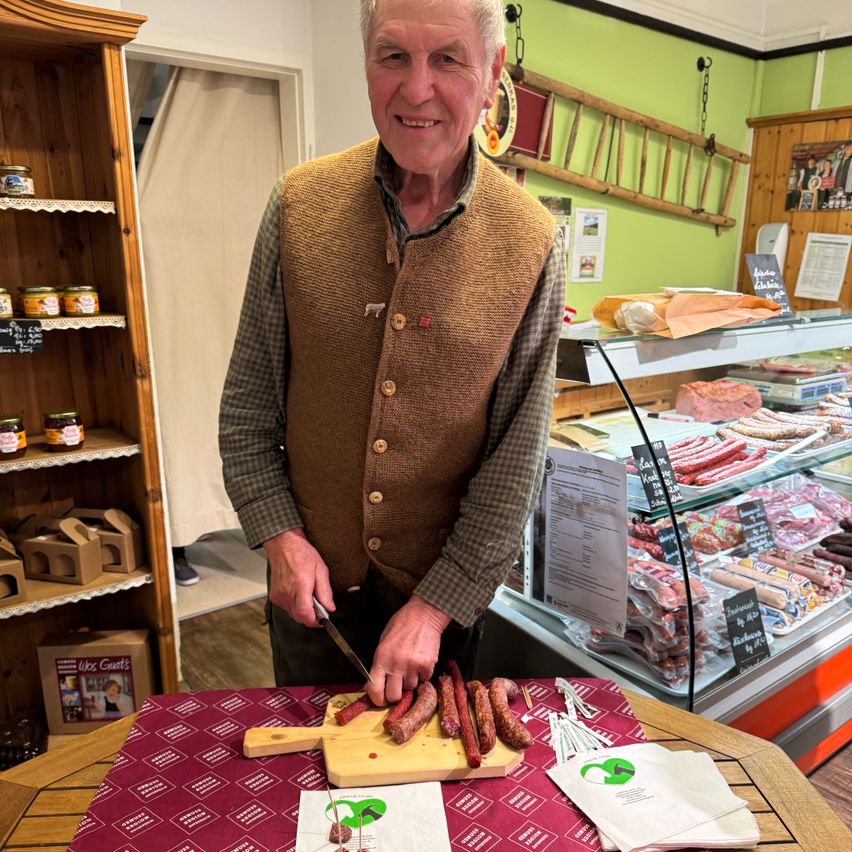 Ein älterer Mann schneidet Wurst auf einem Schneidebrett in einer Metzgerei. Ein Holzregal mit Gläsern ist im Hintergrund.