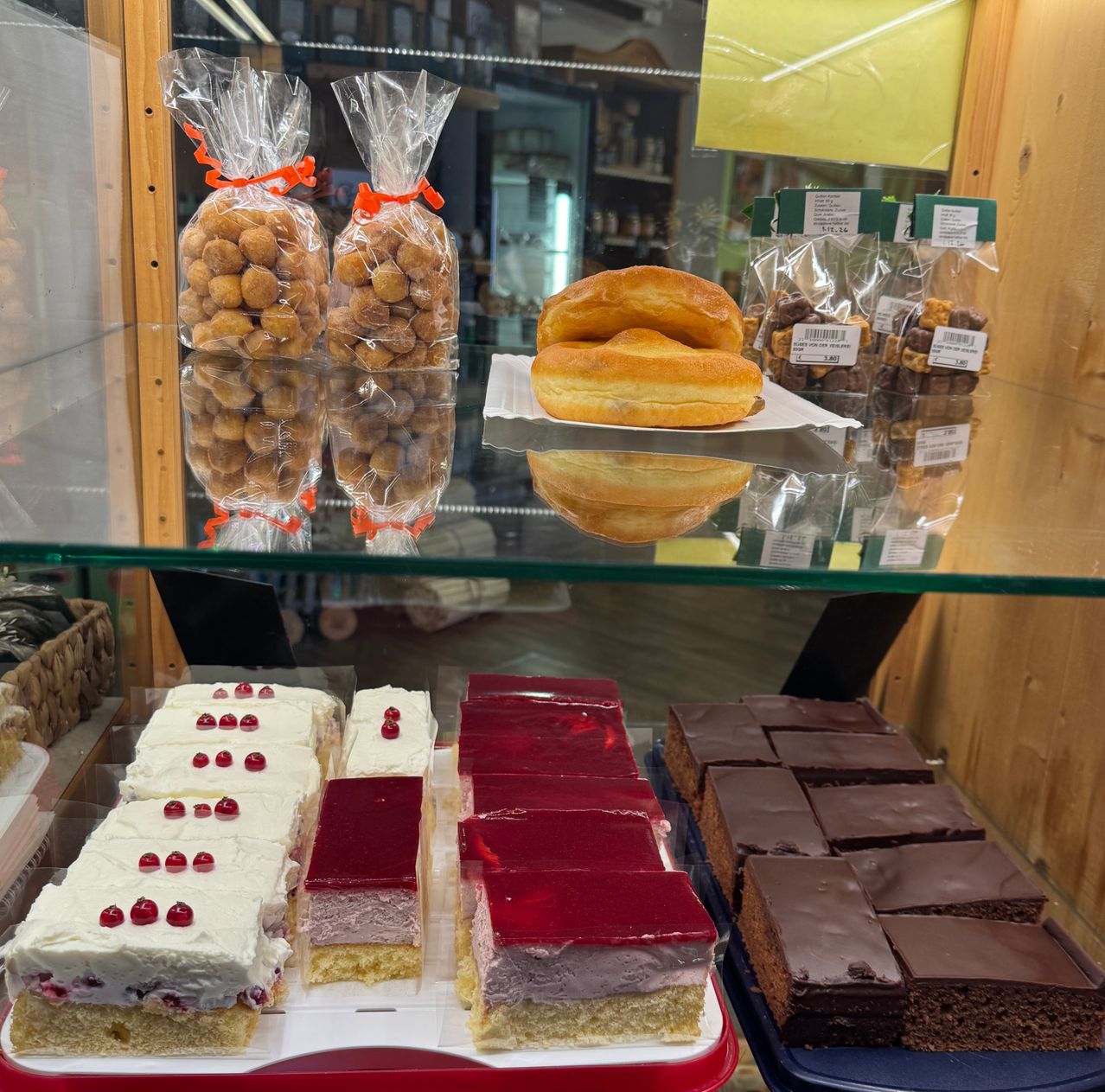 Ein Glasvitrine in einer Bäckerei zeigt eine Auswahl an Desserts, darunter glasierte Donuts, mehrschichtige Kuchen mit roter und weißer Glasur und Schokoladen-Brownies.