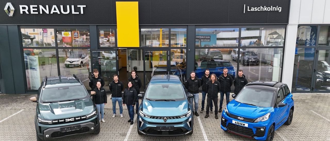 Eine Gruppe von Menschen in schwarzen Jacken steht vor einem Autohaus mit drei Autos, darunter ein blauer Renault und ein blauer Ligier, vor einem Gebäude mit dem Wort Renault darauf.