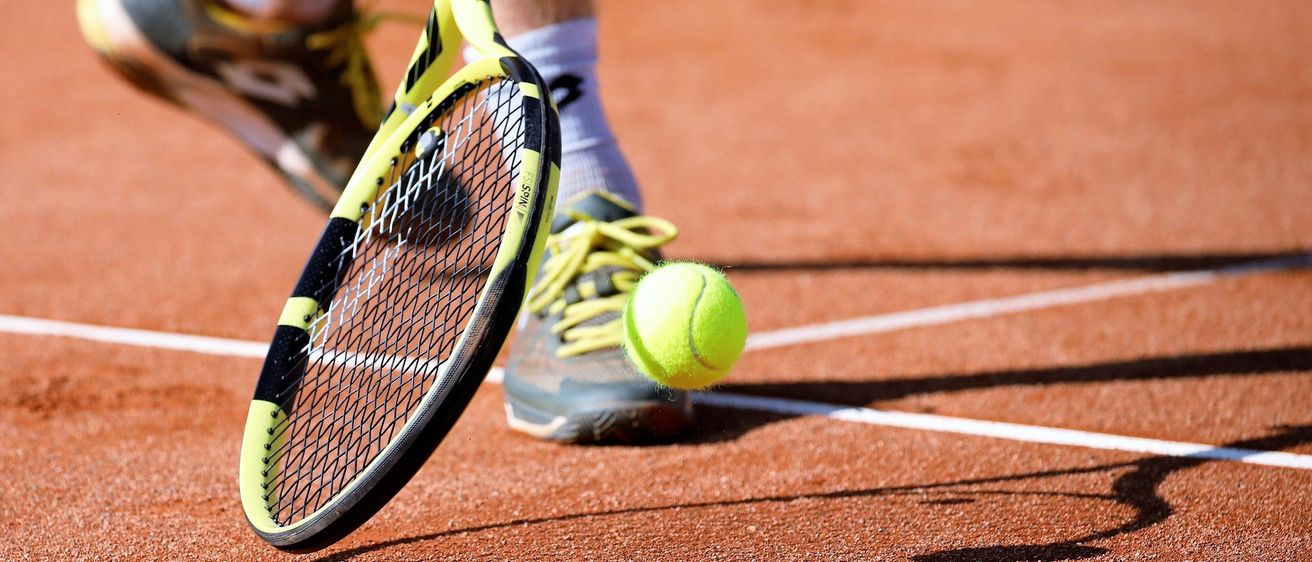 Der Fuß eines Tennisspielers steht auf einem Tennisplatz, mit einem Tennisschläger und einem Ball im Vordergrund.