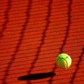 Ein Tennisball liegt auf einem roten Sandplatz, mit Schatten des Netzes auf dem Boden.