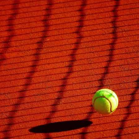 Ein Tennisball liegt auf einem roten Sandplatz, mit Schatten des Netzes auf dem Boden.