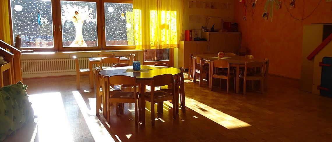 Ein Klassenzimmer mit Holztischen und Stühlen. Die Fenster sind mit gelben Vorhängen verhängt. Ein Heizkörper befindet sich an der Wand, und es gibt einen Schrank mit einigen Gegenständen.