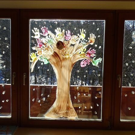 Ein hölzerne Fensterscheibe mit einem Baumdesign, das mit Handabdrücken, Schneeflocken und Lichtern bedeckt ist. Der Baum ist mit farbenfrohen Handabdrücken verziert und von einem verschneiten Hintergrund umgeben.